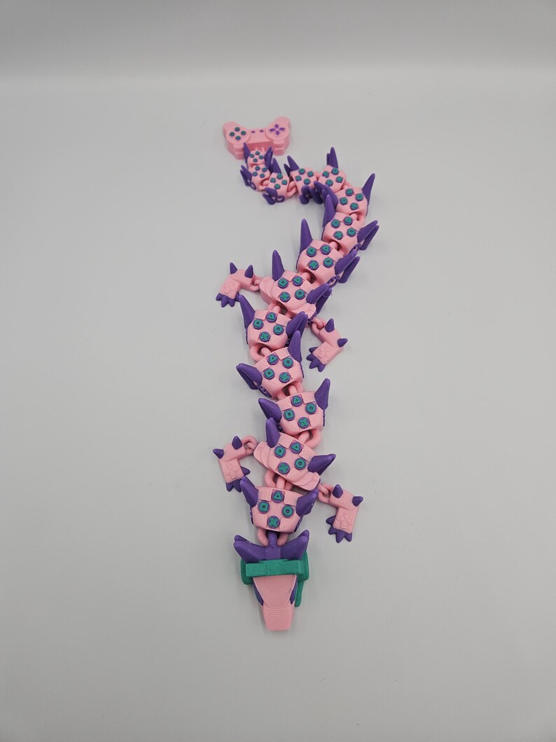 May include: A pink and purple articulated dragon toy with teal accents. The dragon has a controller at the top and a green and pink base. The toy is made of plastic and is designed to be flexible and poseable.