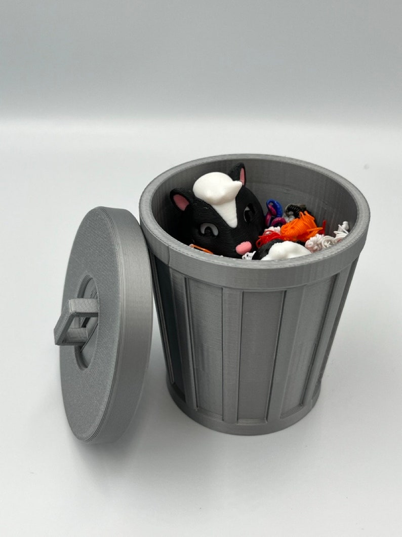 May include: A gray 3D-printed trash can with a lid, containing a black and white skunk figurine and colorful debris. The lid is slightly ajar, revealing the contents. The trash can has a ribbed design.
