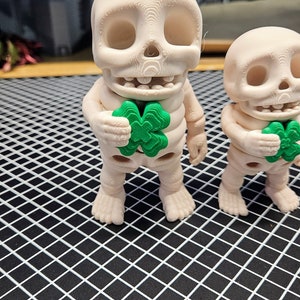 May include: Two white skeleton figurines holding green four-leaf clover charms. The figurines are standing on a black grid surface.