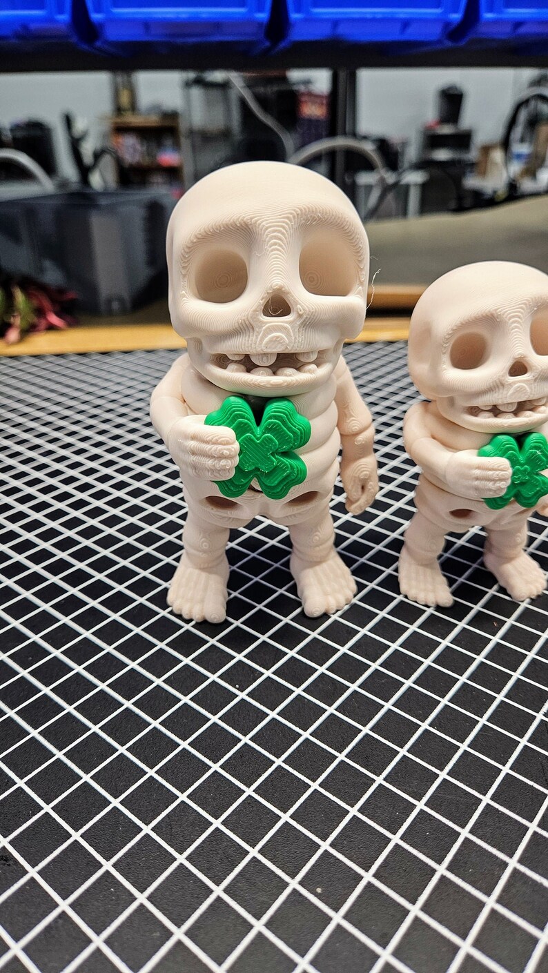 May include: Two white skeleton figurines holding green four-leaf clover charms. The figurines are standing on a black grid surface.
