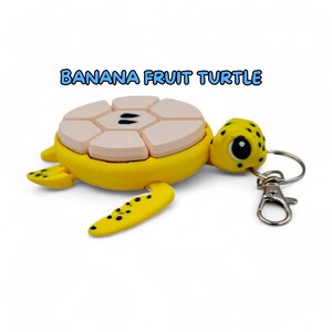 May include: A yellow turtle keychain with a pink and white banana fruit design on its shell. The keychain has a silver metal clasp.