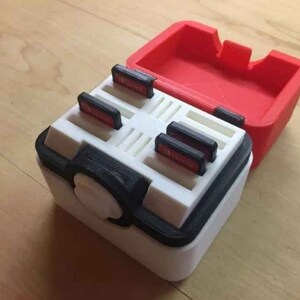 May include: A white and black plastic case with a red lid, designed to hold game cartridges. The case has a circular button on the front and slots for multiple cartridges.