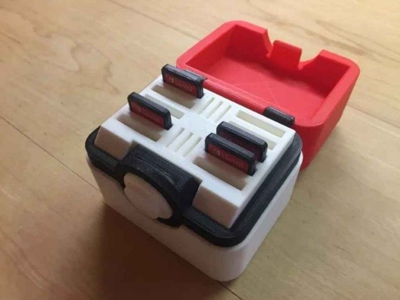 May include: A white and black plastic case with a red lid, designed to hold game cartridges. The case has a circular button on the front and slots for multiple cartridges.