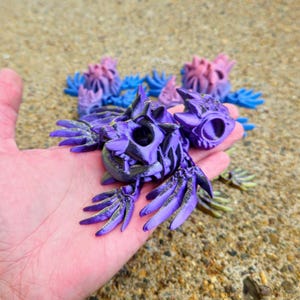 Peut inclure: Un cr&acirc;ne imprim&eacute; en 3D de couleur violette et dor&eacute;e avec des ailes. Le cr&acirc;ne a un trou sur le dessus et est d&eacute;taill&eacute; avec des motifs complexes.