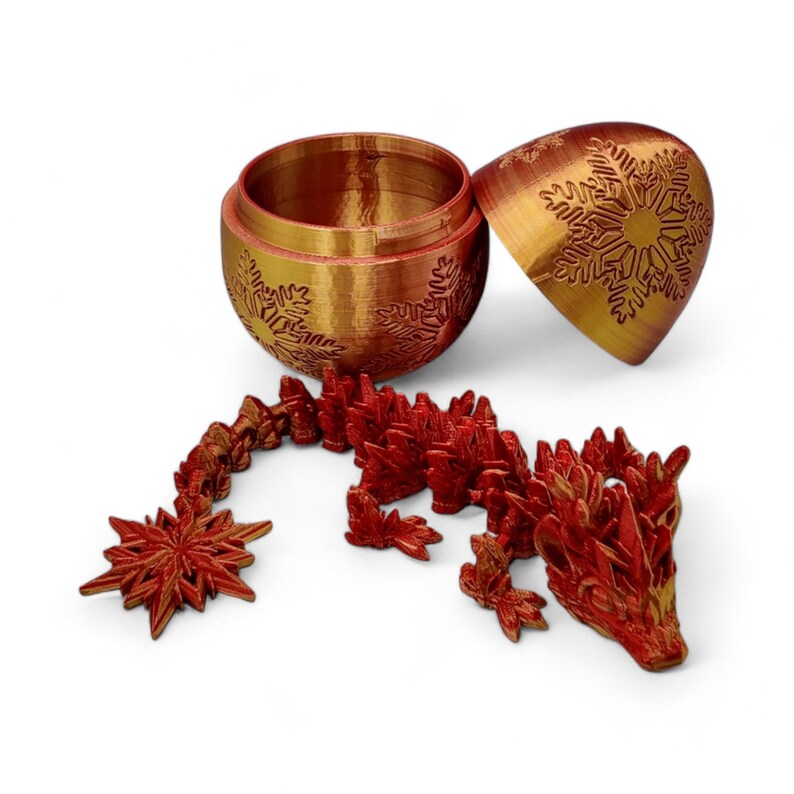 K&ouml;nnte beinhalten: Eine rote und goldene Drachenfigur mit Schneeflocken-Details. Der Drache ist aus Kunststoff und etwa 15 cm lang. Der Drache sitzt auf einem wei&szlig;en Hintergrund. Der Drache ist Teil eines Sets, das eine goldene und rote Kugel enth&auml;lt.