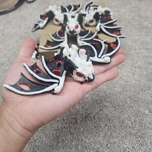 May include: A collection of white, black, and red dragon figurines. The dragons have detailed wings and red eyes. One figurine is held in a hand, while others are arranged on a cardboard base. The figurines are likely 3D printed.