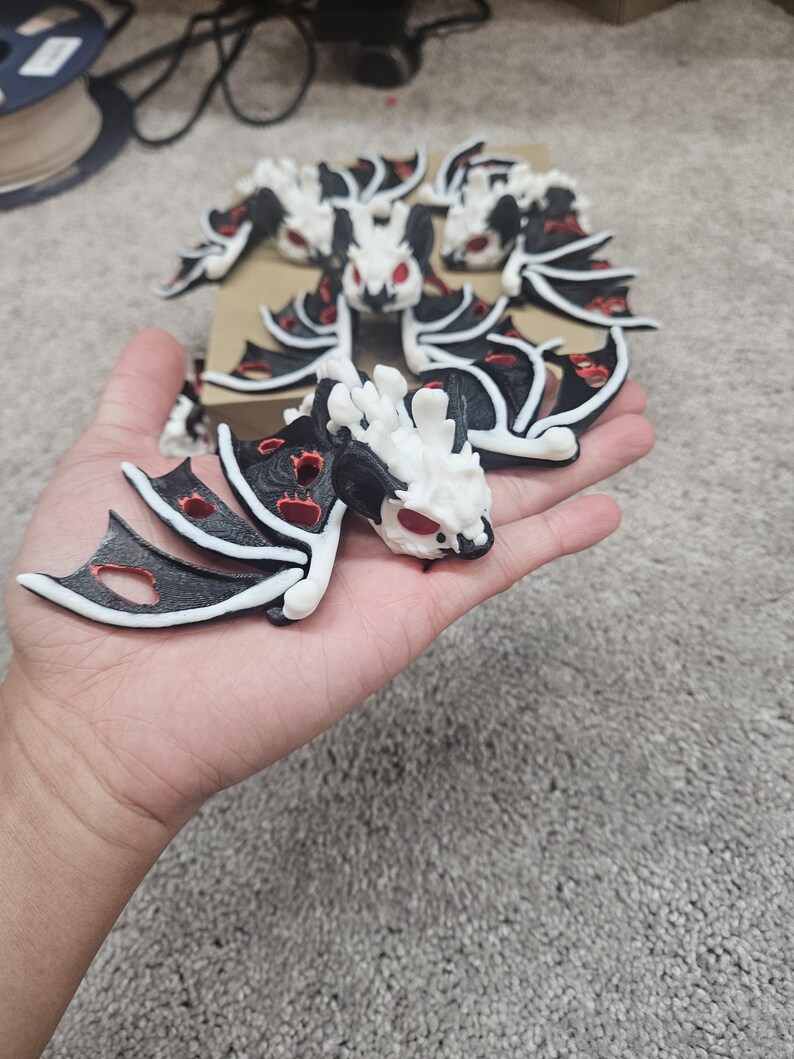May include: A collection of white, black, and red dragon figurines. The dragons have detailed wings and red eyes. One figurine is held in a hand, while others are arranged on a cardboard base. The figurines are likely 3D printed.