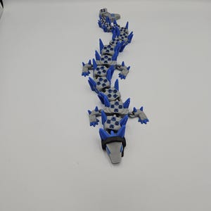 May include: A gray and blue articulated dragon toy. The dragon has a gray head with a black band and blue spikes along its back. The body is made of interlocking pieces, allowing for flexible movement. The toy is designed to resemble a dragon.