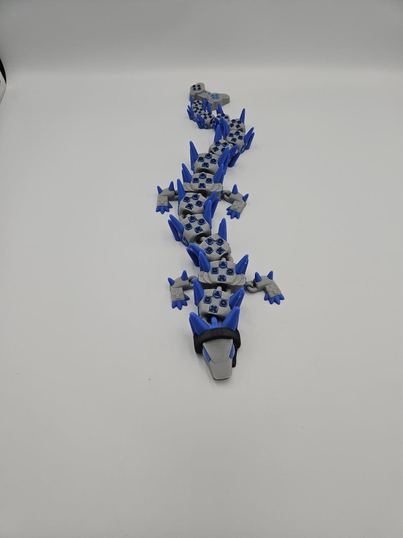 May include: A gray and blue articulated dragon toy. The dragon has a gray head with a black band and blue spikes along its back. The body is made of interlocking pieces, allowing for flexible movement. The toy is designed to resemble a dragon.