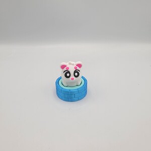 May include: A white and pink cartoon animal figure with large black eyes and pink accents, sitting on a blue, brick-patterned base. The figure has small pink ears and a round, white body.