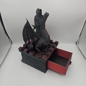 May include: A black and red dragon figurine atop a castle-like box with a drawer. The dragon has its mouth open and wings spread. The drawer is red inside. The box is black with red accents.