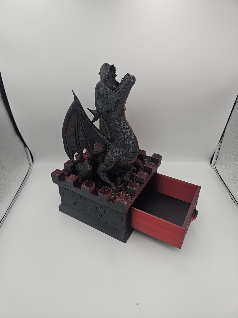 May include: A black and red dragon figurine atop a castle-like box with a drawer. The dragon has its mouth open and wings spread. The drawer is red inside. The box is black with red accents.