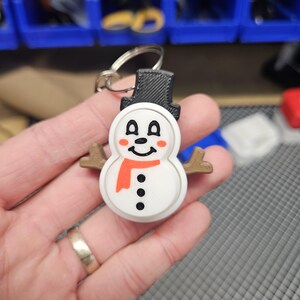 Pode incluir: Um chaveiro de boneco de neve branco com um chap&eacute;u preto, uma echarpe laranja e olhos e bot&otilde;es pretos. O boneco de neve tem bra&ccedil;os de galho castanho e uma cara sorridente.