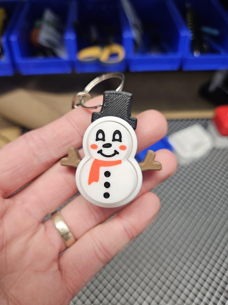 Pode incluir: Um chaveiro de boneco de neve branco com um chap&eacute;u preto, uma echarpe laranja e olhos e bot&otilde;es pretos. O boneco de neve tem bra&ccedil;os de galho castanho e uma cara sorridente.