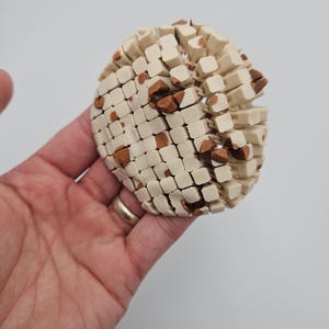 May include: A spherical, off-white object with a mosaic pattern of small, square blocks and scattered brown accents. The object is held in a hand against a plain background, showcasing its textured surface and unique design.
