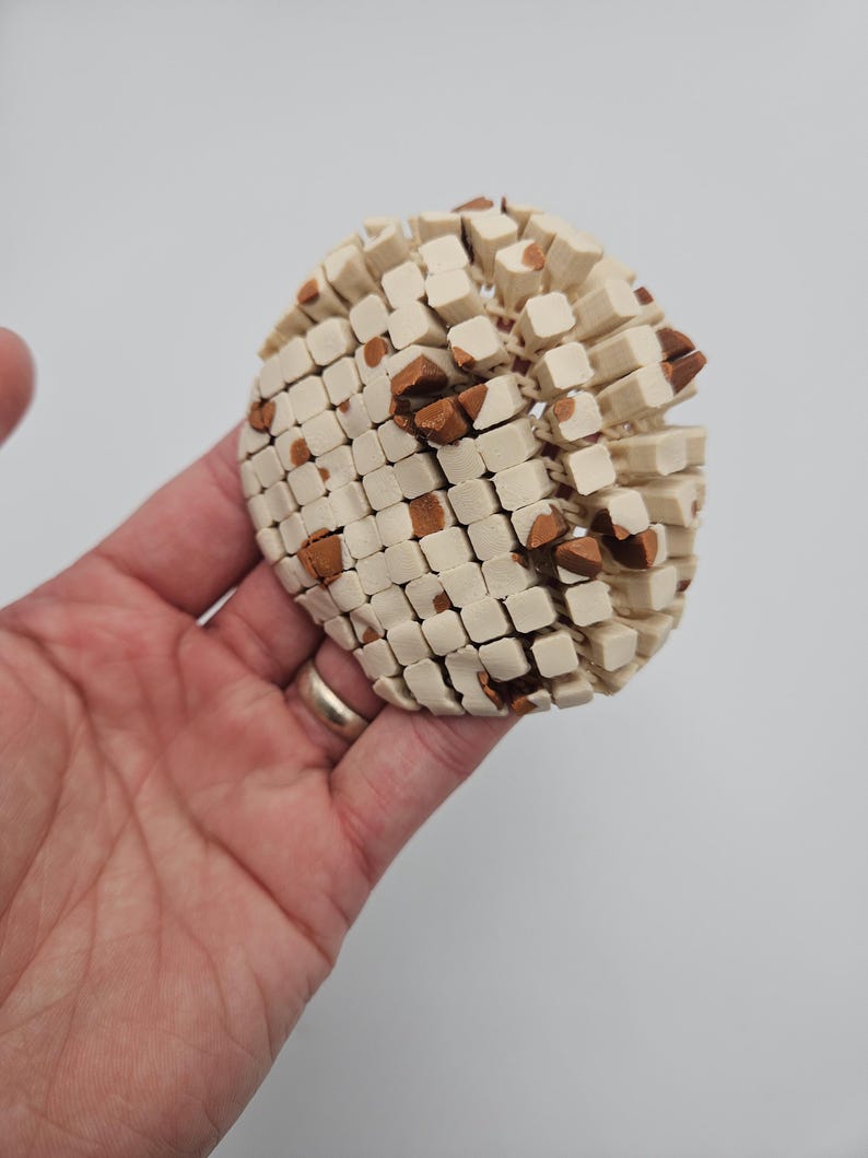 May include: A spherical, off-white object with a mosaic pattern of small, square blocks and scattered brown accents. The object is held in a hand against a plain background, showcasing its textured surface and unique design.