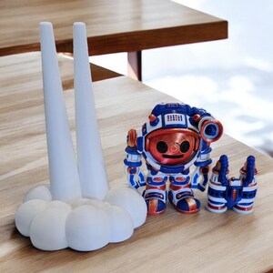 May include: A white ceramic rocket ship with two tall, pointed towers and a cloud-shaped base. A small, blue and red astronaut figurine with a copper helmet stands in front of the rocket.