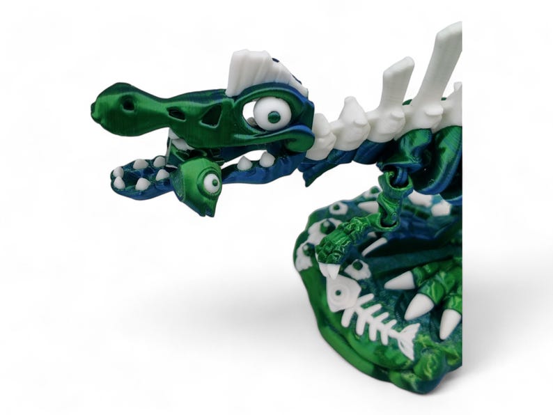 May include: A green and white plastic dinosaur toy with a skeleton design. The dinosaur has large eyes and is holding a fish in its mouth.