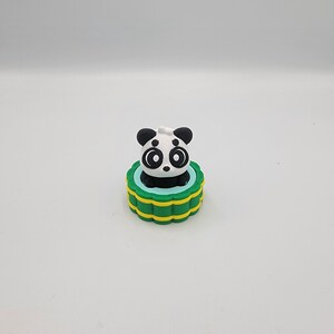 May include: A panda figurine with a black and white face and ears sits atop a green and yellow base. The panda has large, round eyes and a small black nose. The base has a blue interior.