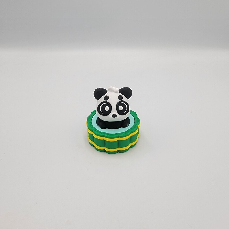 May include: A panda figurine with a black and white face and ears sits atop a green and yellow base. The panda has large, round eyes and a small black nose. The base has a blue interior.