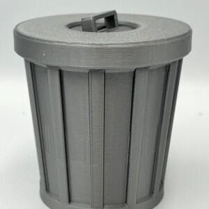 May include: A gray, miniature trash can with a lid. The can has vertical lines and a handle on the lid. The design is simple and modern, suitable for a desk or small space. The can is made of a solid, matte material.