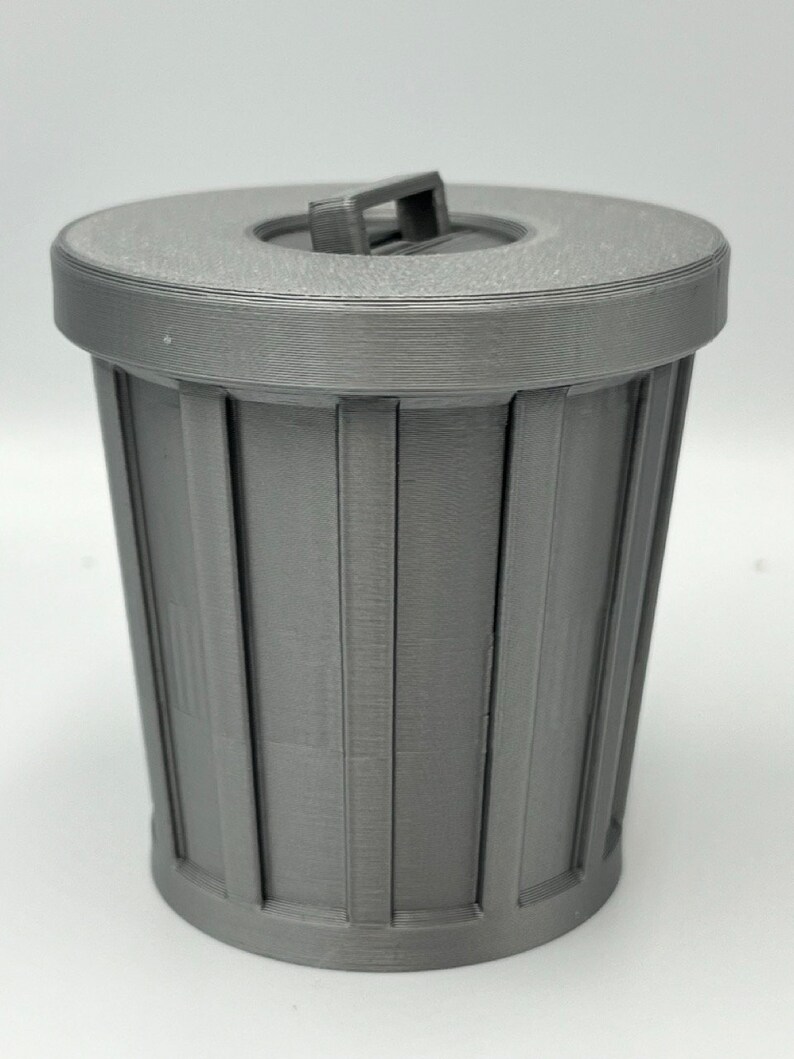 May include: A gray, miniature trash can with a lid. The can has vertical lines and a handle on the lid. The design is simple and modern, suitable for a desk or small space. The can is made of a solid, matte material.