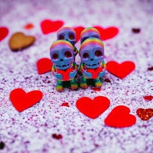 May include: Four colourful skeleton figurines holding red hearts, surrounded by red hearts on a white background with pink and purple speckles.