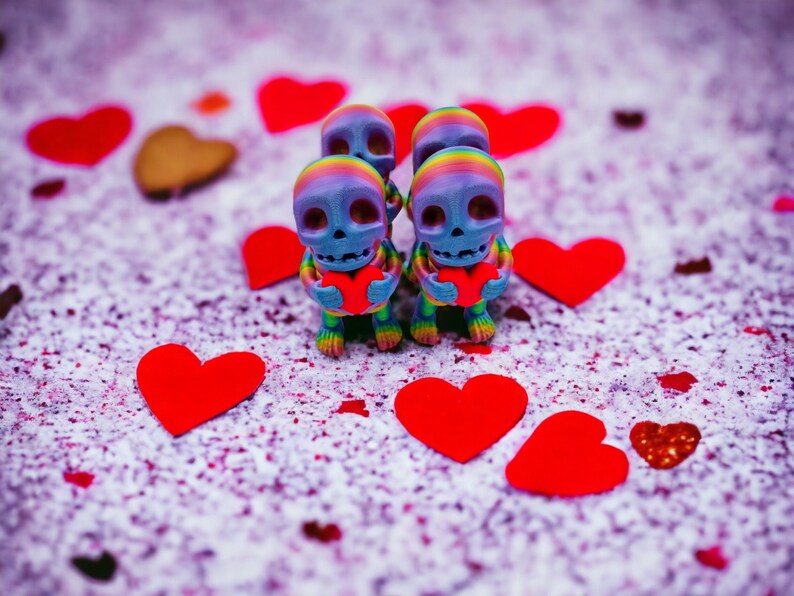 May include: Four colourful skeleton figurines holding red hearts, surrounded by red hearts on a white background with pink and purple speckles.