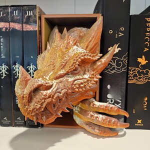 May include: A detailed, bronze-colored dragon head bookend. The dragon's head and claw extend from a wooden box, with sharp teeth and textured scales. The bookend is designed to hold books upright on a shelf.