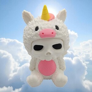 Puede incluir: Una figura blanca de una calavera con un disfraz de unicornio esponjoso con un cuerno amarillo y detalles en rosa. La figura tiene ojos negros y un c&iacute;rculo rosa en el pecho. El fondo es un cielo azul con nubes blancas.