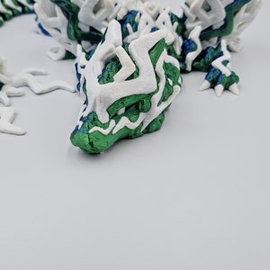 May include: A 3D printed green and white dragon figurine with intricate details. The dragon has a long tail and large wings.