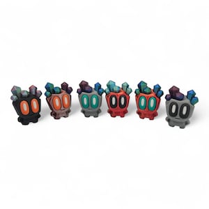 May include: Six colourful, stylised figurines with crystal-like headpieces. Each figure has a unique colour scheme, including black, orange, green, red, and grey. The figurines have large, round eyes and a small, smiling mouth.