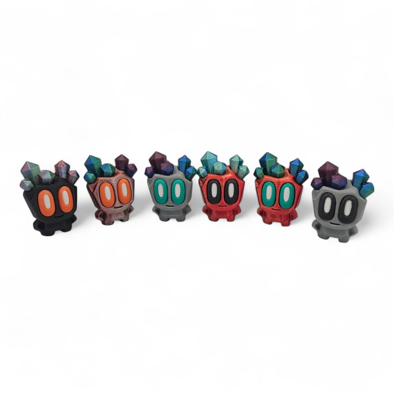 May include: Six colourful, stylised figurines with crystal-like headpieces. Each figure has a unique colour scheme, including black, orange, green, red, and grey. The figurines have large, round eyes and a small, smiling mouth.