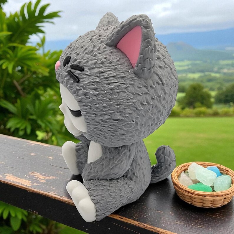 May include: A gray cat figurine with pink inner ears and white paws sits on a wooden surface. The cat is wearing a hood and has black details for the eyes and nose. A small woven basket with colorful stones sits nearby. The background shows a green landscape.