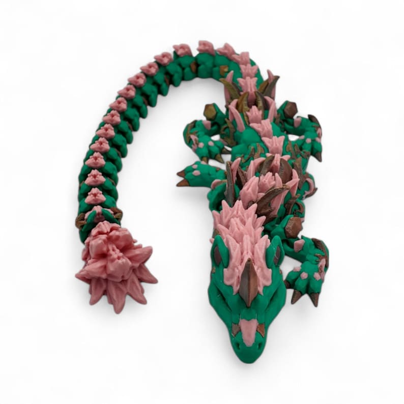 May include: A handcrafted, whimsical dragon figurine in shades of green and pink. The dragon's body is a vibrant green, with pink accents along its spine and head. The dragon's tail curves gracefully, and the head is detailed with intricate features.