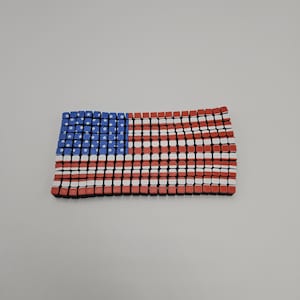 May include: A rectangular American flag design composed of small, colorful blocks. The flag features a blue field with white stars in the upper left corner and red and white stripes across the rest of the flag.