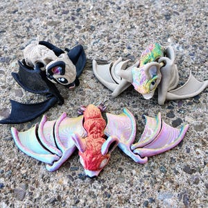 May include: Three small bat figurines with wings outstretched. One is black, one is beige with brown accents, and one is red with iridescent wings.