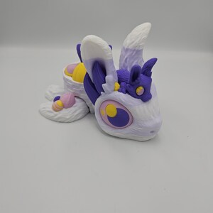 Pode incluir: Uma criatura de fantasia impressa em 3D em tons de branco, roxo, amarelo e rosa. A criatura tem olhos grandes, orelhas fofas e um pequeno companheiro. O design lembra um drag&atilde;o ou besta m&iacute;tica.