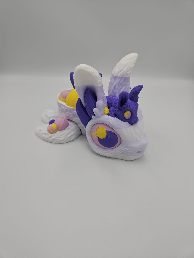 Pode incluir: Uma criatura de fantasia impressa em 3D em tons de branco, roxo, amarelo e rosa. A criatura tem olhos grandes, orelhas fofas e um pequeno companheiro. O design lembra um drag&atilde;o ou besta m&iacute;tica.