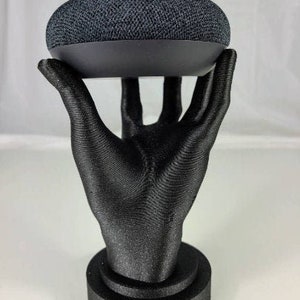 Op de afbeelding: Een zwarte 3D-geprinte hand die een zwarte Google Home Mini smart speaker vasthoudt. De hand staat op een zwarte basis.