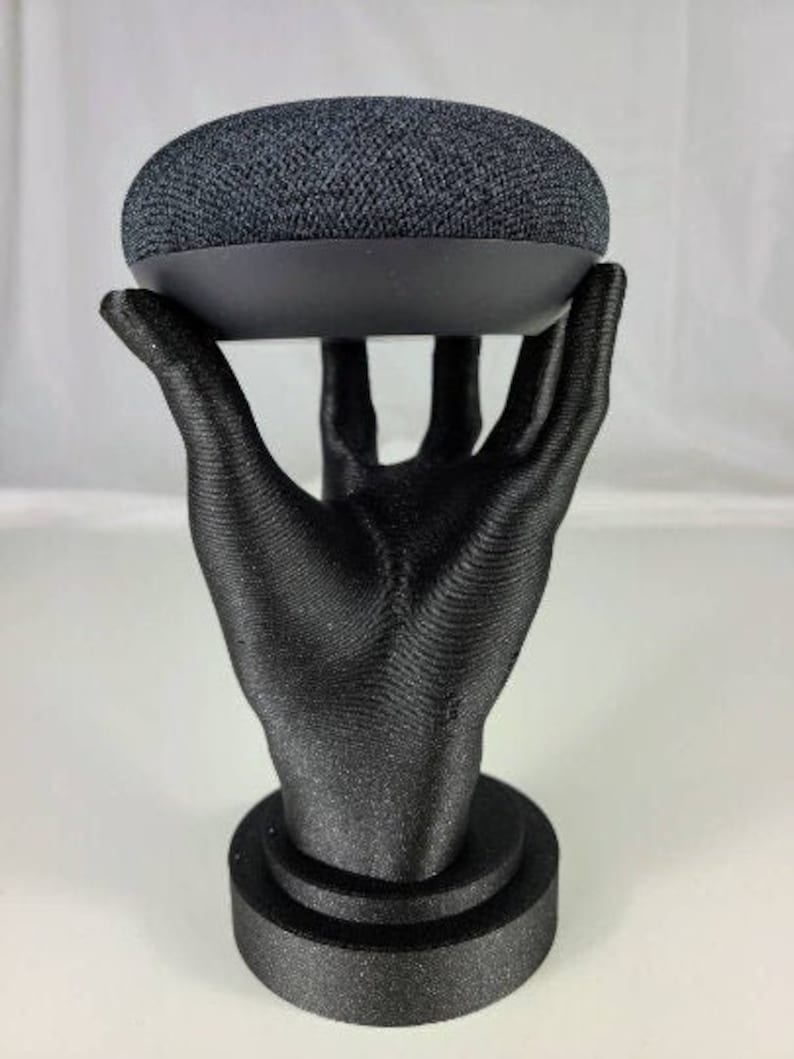 Op de afbeelding: Een zwarte 3D-geprinte hand die een zwarte Google Home Mini smart speaker vasthoudt. De hand staat op een zwarte basis.