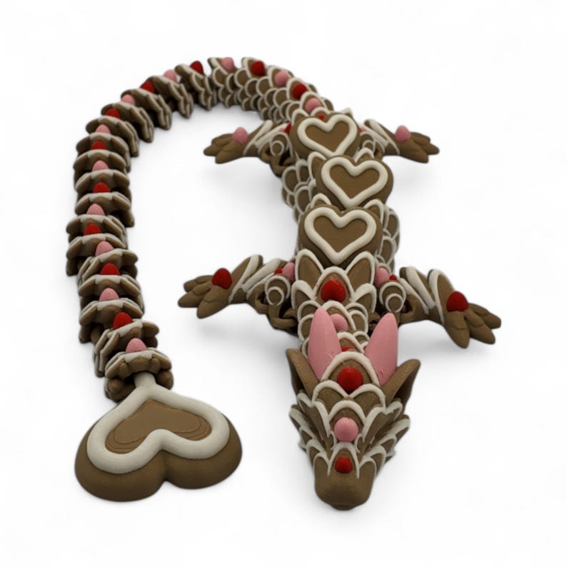 May include: A brown and white dragon figurine with heart-shaped accents and pink details. The dragon's body is segmented, with a curved tail and a detailed head. The design incorporates red and white embellishments, creating a whimsical and decorative appearance.