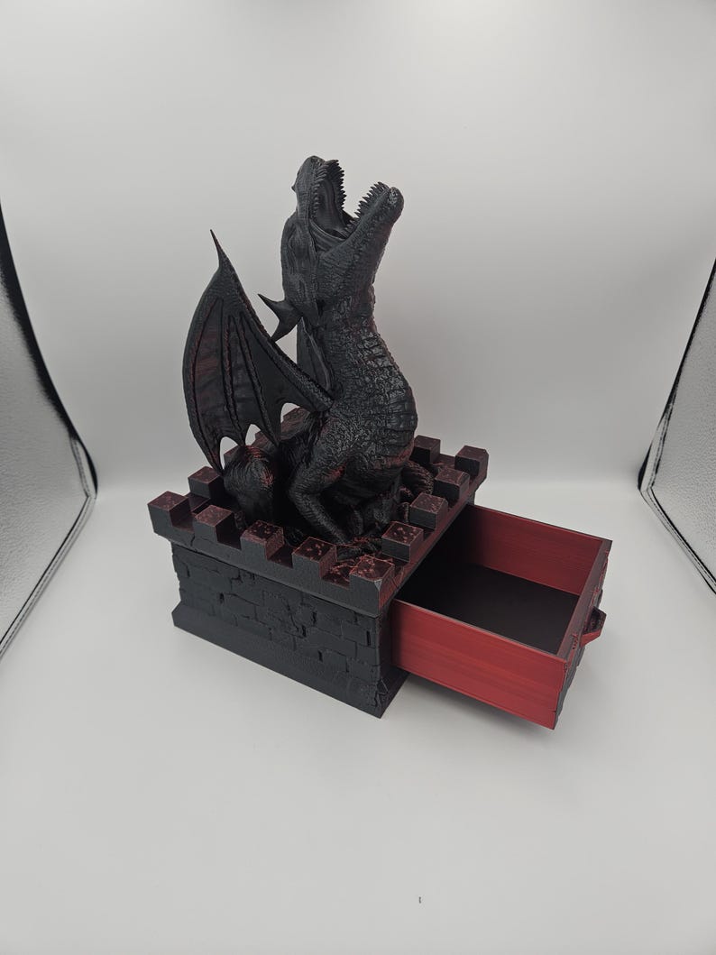 May include: A black dragon figurine with open mouth and detailed scales sits atop a black and red box with an open drawer. The dragon has large wings and is posed as if roaring. The box has a castle-like design.