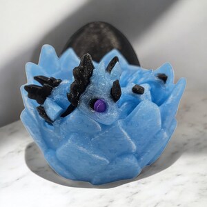 May include: A small, blue, 3D printed dragon sculpture with black wings and purple eyes, nestled inside a blue, 3D printed egg.