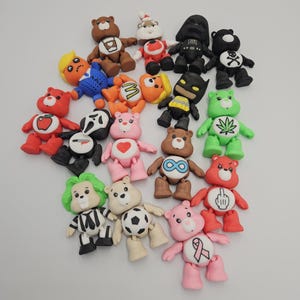 May include: A collection of colorful bear figurines, each uniquely designed. The bears feature various themes, including a Santa Claus, a Batman, and a bear with a cannabis leaf. The figurines are made of a smooth material and are arranged on a white surface.