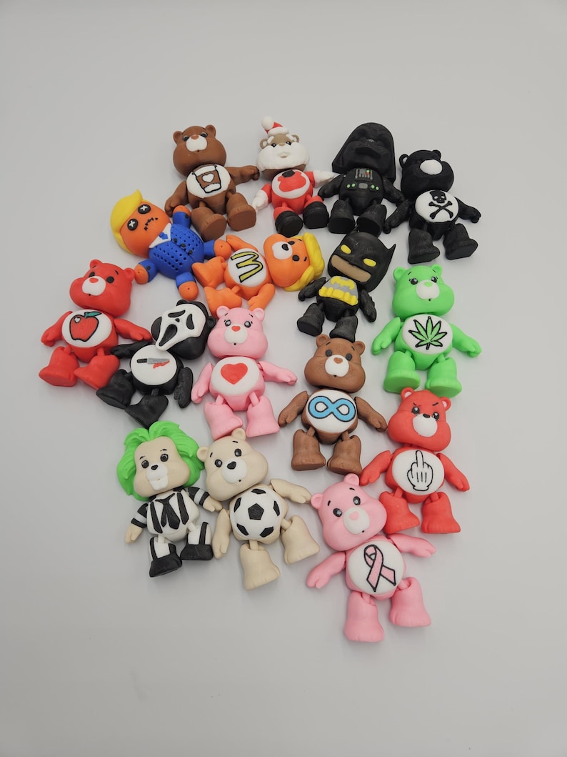 May include: A collection of colorful bear figurines, each uniquely designed. The bears feature various themes, including a Santa Claus, a Batman, and a bear with a cannabis leaf. The figurines are made of a smooth material and are arranged on a white surface.