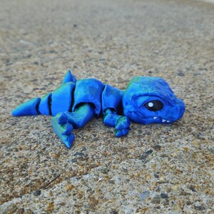 May include: A small, blue and green, articulated dinosaur toy. The dinosaur is lying on its side with its head turned to the side. The toy has a detailed face with a small, black eye.