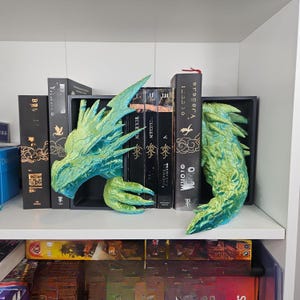 May include: A teal and green dragon head and tail bookends are displayed on a white shelf. The bookends are placed on either side of a row of black books. The dragon head is on the left, and the tail is on the right.