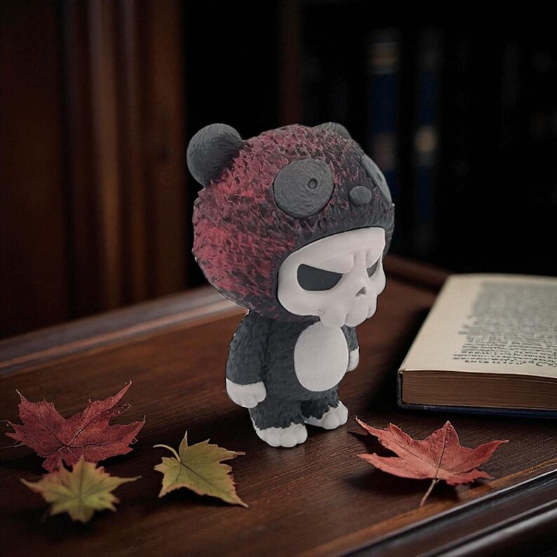 May include: A figurine of a panda wearing a skull mask and a panda suit. The panda suit is black and white, and the mask is white with black eyes. The panda's hat is red and black. The figurine is on a wooden surface with autumn leaves.
