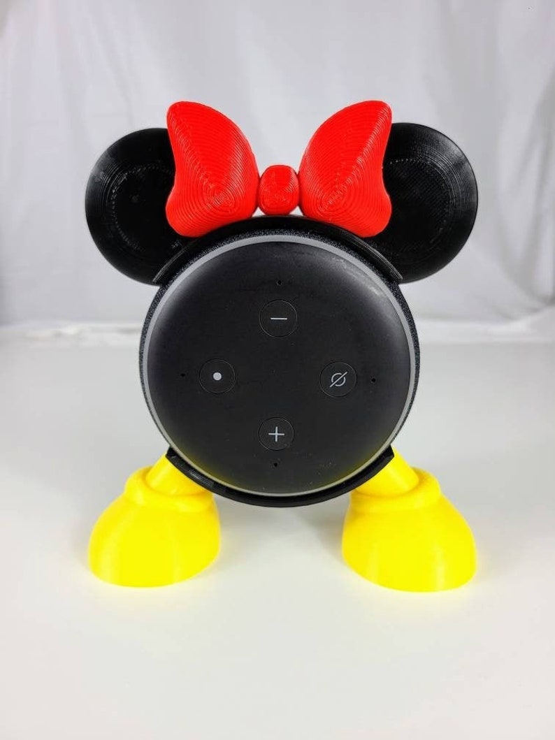 Puede incluir: Un soporte para altavoz inteligente impreso en 3D de color negro y amarillo, dise&ntilde;ado para parecerse a Mickey Mouse. El soporte tiene un lazo rojo en la parte superior y pies amarillos.